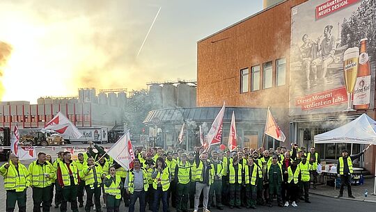 Große Gruppe Streikender in neongelben Warnwesten mit Fahnen und Pyrotechnik vor der Brauerei Berliner Kindl. 